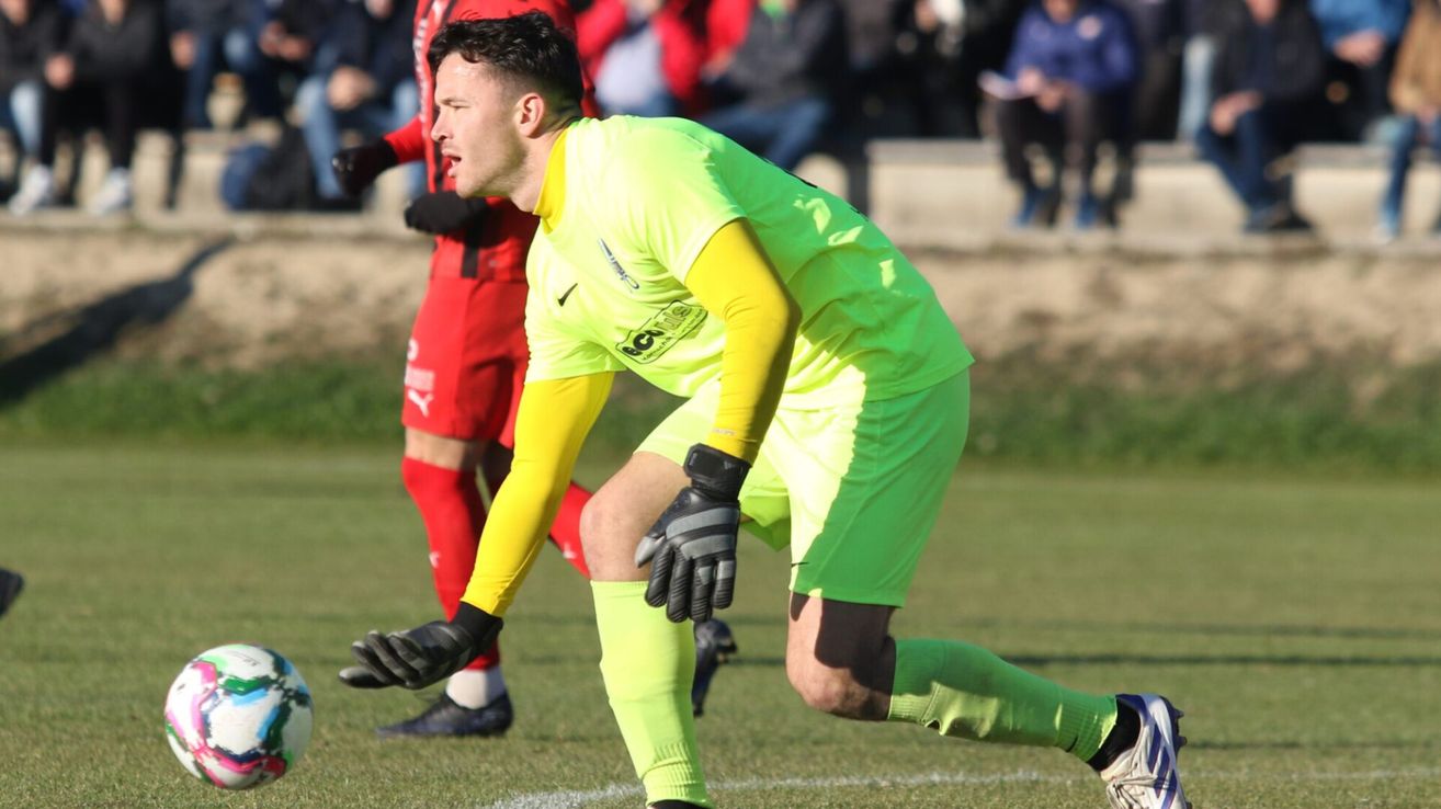 Ein Fußballspieler in einem grünen Trikot beugt sich während eines Spiels an einem sonnigen Tag. Im unscharfen Hintergrund sind Zuschauer und ein Rasenfeld zu sehen.