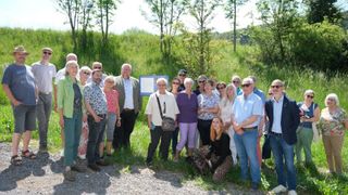 Eine Gruppe von Menschen in Freizeitkleidung posiert für ein Foto im Freien, mit einem Schild im Hintergrund. Sie sind von üppiger Vegetation umgeben, einschließlich Bäumen und Gras.