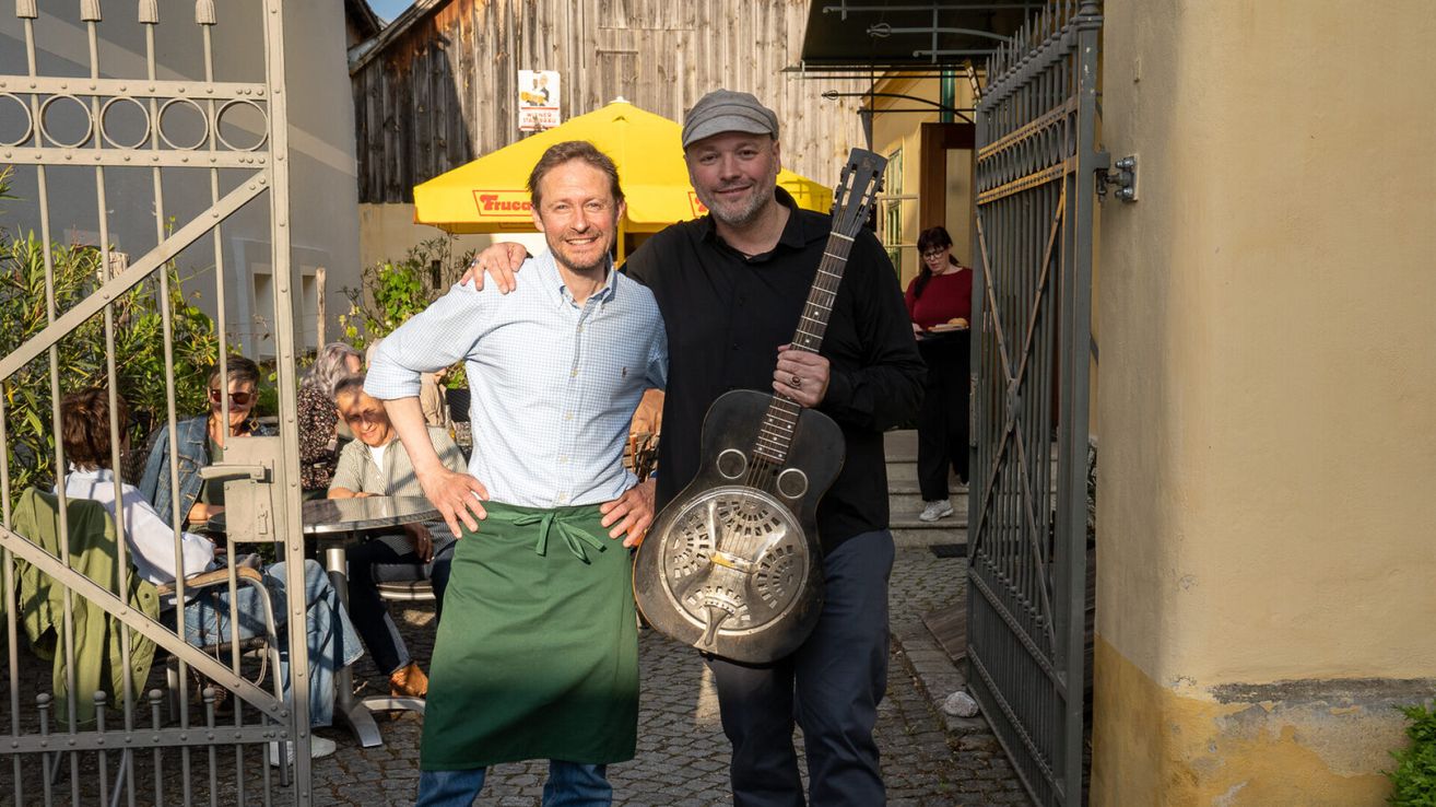 Zwei Männer stehen vor einem Gebäude mit einem gelben Schirm, einer trägt eine grüne Schürze und hält eine Gitarre. Hinter ihnen steht eine Frau und ein Gebäudeeingang.