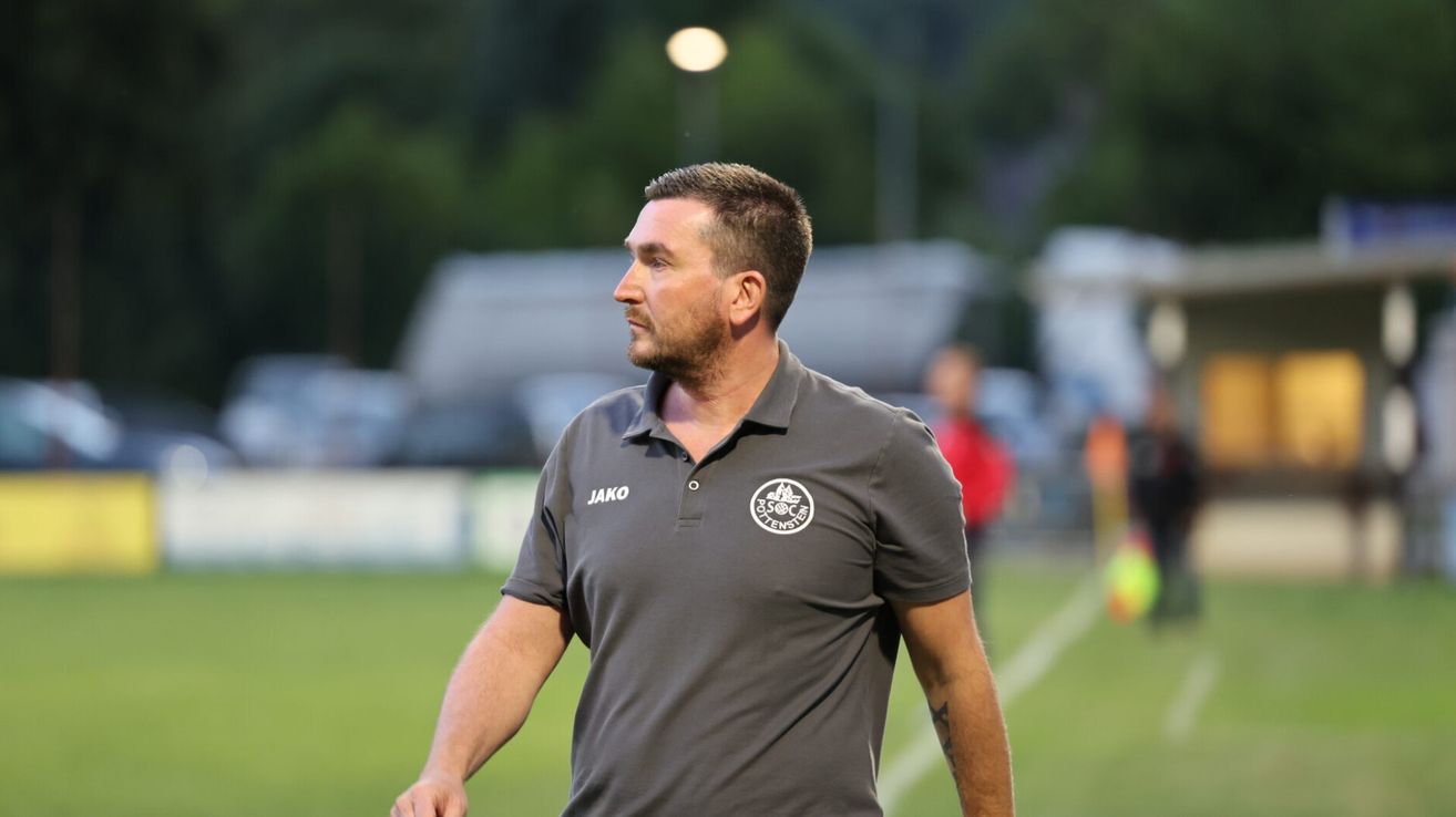 A man wearing a dark gray polo shirt with the word Jako on the left side and a logo on the right side is walking on a football field at night.