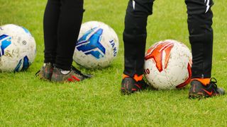 Zwei Personen stehen auf einem Feld mit Fußballen. Ein Ball hat ein blaues Design, der andere ein rotes und weißes Design. Beide Spieler tragen schwarze Hosen.