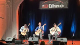 Drei Männer spielen Gitarren auf der Bühne, mit einem blauen Vorhang im Hintergrund und dem Wort 'Bühne' darüber. Das Publikum sitzt vor ihnen.
