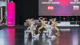 Fünf junge Frauen tanzen auf der Bühne in goldenen Paillettenkleidern. Im Hintergrund zeigt eine Leinwand '108 AUT DANCING QUEENS'.