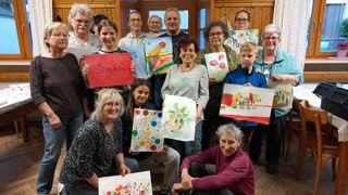 Eine Gruppe Erwachsener und Kinder posiert drinnen mit Kunstwerken. Eine Frau hält ein Schild mit Sophia. Ein Kind hält ein Bild mit Blumen. Ein anderes hält ein Gemälde mit einem Hund.