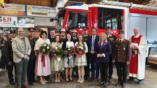 Eine Gruppe von Personen steht vor einem roten Feuerwehrauto. Mehrere Frauen halten Blumensträuße, während Männer in Militäruniformen hinter ihnen stehen.