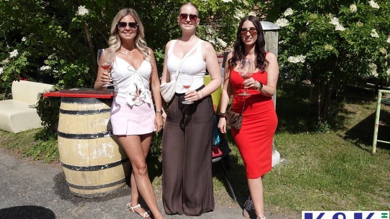 Drei Frauen in Sommerkleidung posieren für ein Foto. Eine hält ein Weinglas, eine andere trägt ein weißes Kleid und die dritte ein rotes Kleid. Ein Holzfass und Grünpflanzen sind im Hintergrund zu sehen.