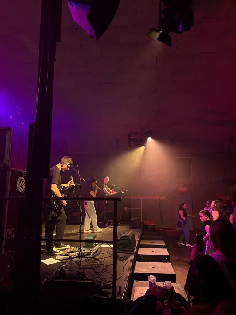Eine Band spielt auf der Bühne mit einem Scheinwerfer. Der Leadsänger hält ein Mikrofon und der Gitarrist spielt eine E-Gitarre. Das Publikum im Hintergrund schaut zu.
