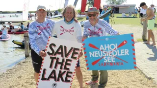 Drei Männer stehen am Strand und halten Schilder mit der Aufschrift 'Rettet den See' und 'AHOI Neusiedler See Lakemania'. Sie tragen gestreifte Hemden und Hüte. Im Hintergrund sind Boote und Menschen auf dem Wasser zu sehen.