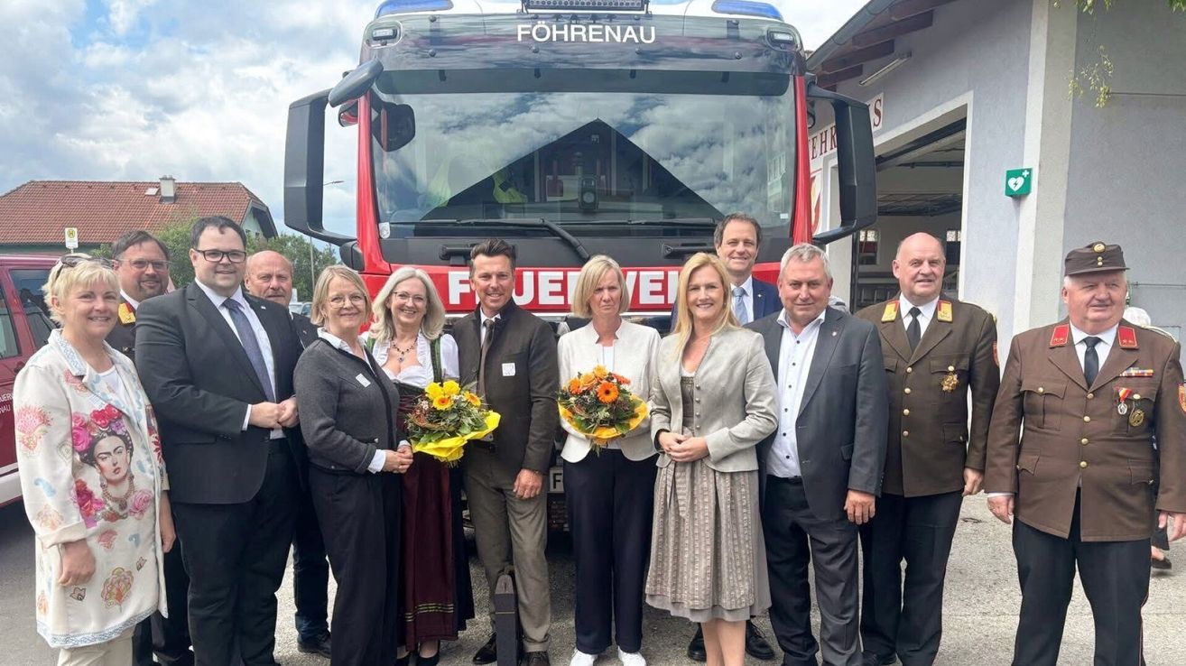 Eine Gruppe von Menschen steht vor einem roten Feuerwehrauto. Sie lächeln und halten Blumen. Links steht eine Frau mit Brille.