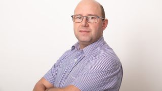 Ein Mann mit Brille steht mit verschränkten Armen. Er trägt ein blau kariertes Poloshirt. Er lächelt und schaut nach vorne.