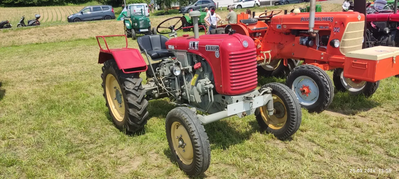 Ein roter Traktor mit schwarzem Sitz steht auf Gras. Andere Fahrzeuge und Menschen sind im Hintergrund.