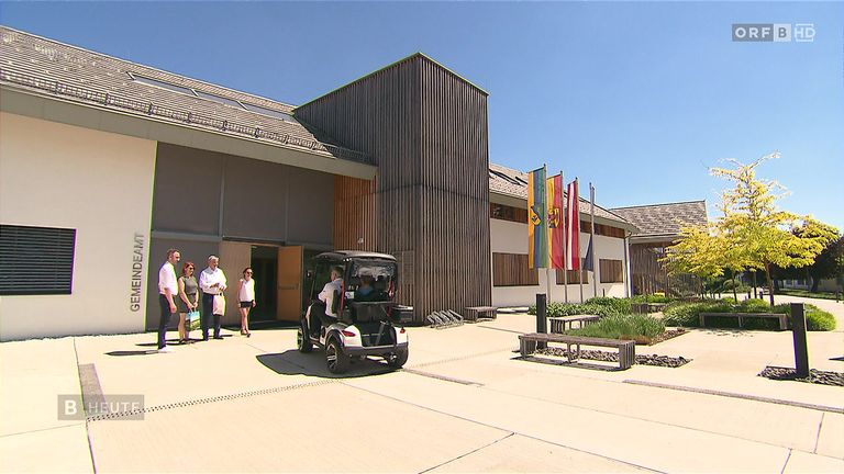 Eine Gruppe von Menschen steht vor einem modernen Gebäude mit Holzakzenten, Fahnen und einem Golfcart im Vordergrund.