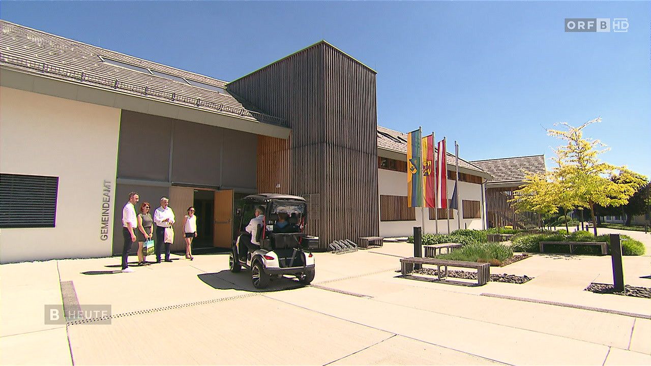 Eine Gruppe von Menschen steht vor einem modernen Gebäude mit Holzakzenten, Fahnen und einem Golfcart im Vordergrund.