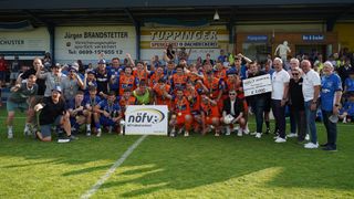 Eine Fußballmannschaft in orangefarbenen Trikots feiert auf einem Feld mit weißer Linie und hält Medaillen und Trophäen. Ein Mann in Weiß hält einen großen Scheck über 3.000 €. Im Hintergrund befindet sich ein Banner mit 'TUPPINGER'.