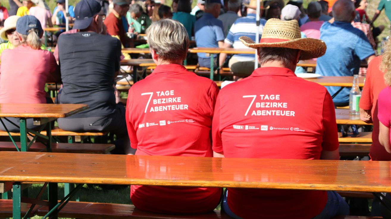 Zwei Personen in roten T-Shirts mit '7 Tage Bezirke Abenteuer' sitzen an einem Holztisch bei einer Outdoor-Veranstaltung.