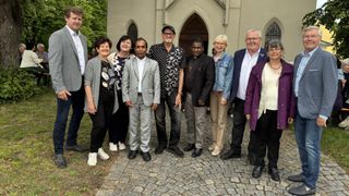 Eine Gruppe von Menschen in formeller Kleidung steht vor einer Kirche. Sie lächeln und posieren für ein Foto. Einige tragen Brillen und Halsketten. Hinter ihnen ist ein Gebäude mit Bogenfenstern zu sehen.