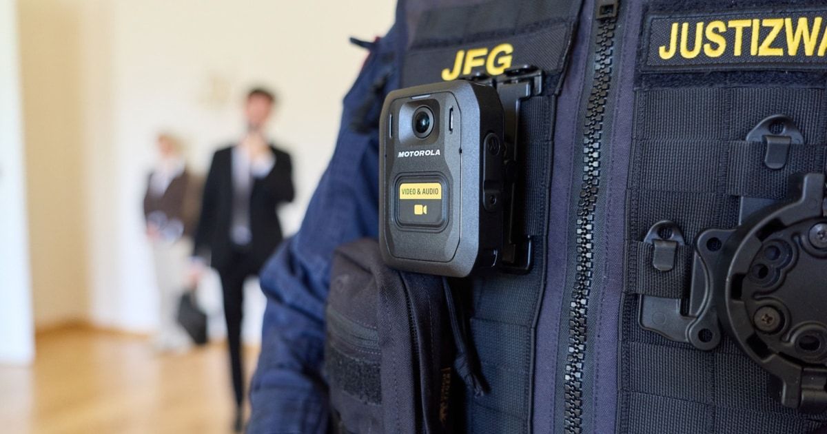 Die Uniform eines Polizisten mit einer Motorola-Körperkamera. Die Kamera hat ein gelbes Etikett, das 'Video & Audio' steht. Der Polizist ist im Hintergrund zu sehen, wie er geht.
