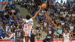 Ein Basketballspieler springt, um den Ball zu werfen, während zwei andere Spieler versuchen, den Schuss zu blockieren. Der Spieler in Weiß und Blau trägt die Nummer 20. Der Spieler in Schwarz und Braun trägt die Nummer 5.