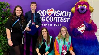 Eine Gruppe von vier Personen vor einem violetten Eurovision 2026-Hintergrund. Ein Maskottchen und ein Mann im Anzug sind ebenfalls anwesend.