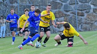 Fünf junge Fußballspieler spielen auf einem Rasenfeld. Ein Spieler in Blau versucht, den Ball zu schießen, während andere versuchen, ihn zu blockieren. Sie tragen Trikots mit verschiedenen Sponsoren.