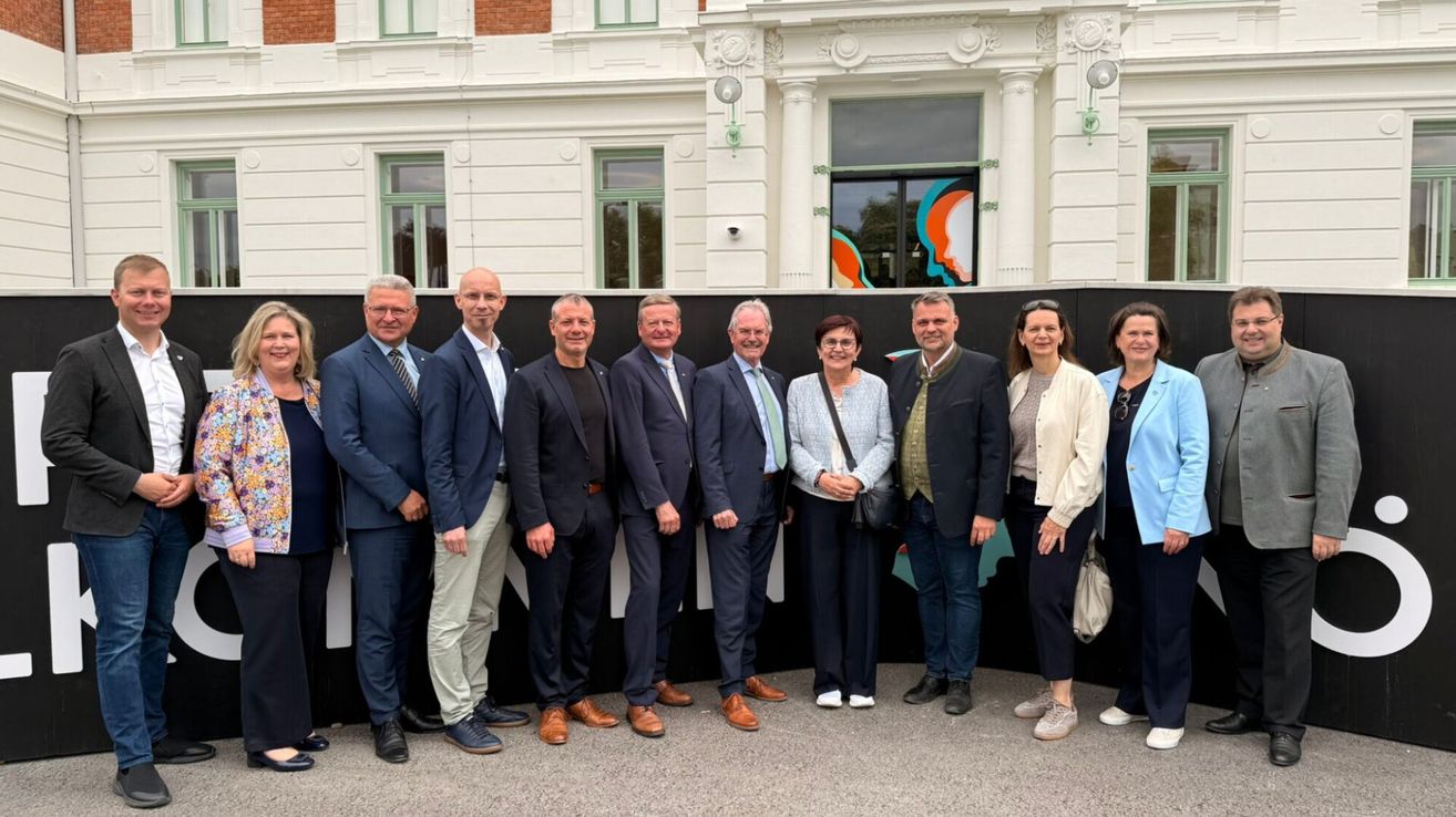 Eine Gruppe von formell gekleideten Personen, einschließlich Männer und Frauen, steht vor einem Gebäude. Das Gebäude hat viele Fenster und ein Banner.