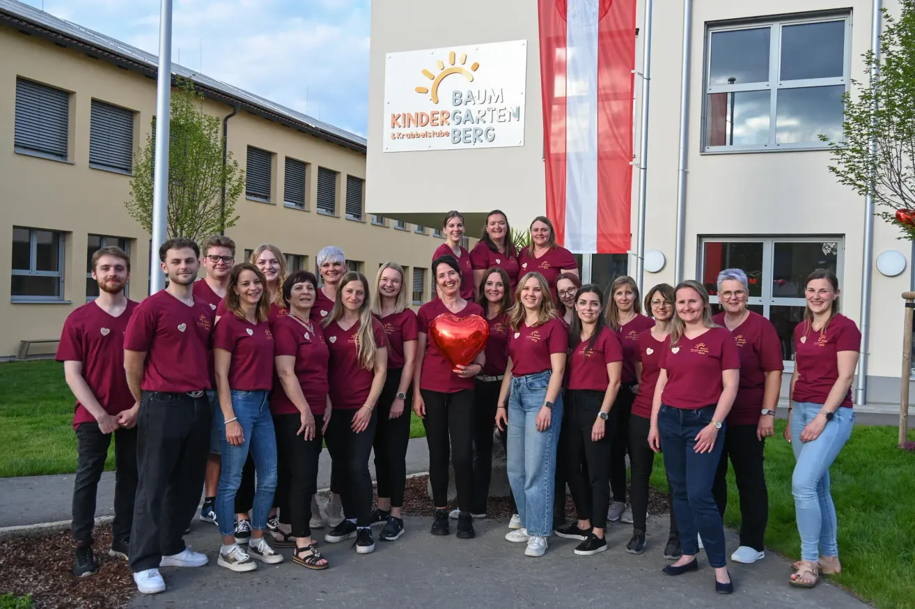 Eine Gruppe von Erwachsenen in Weinroten T-Shirts, die ein rotes Herz halten, stehen vor einem Gebäude mit einem Schild, das Baum Kindergarten Berg sagt.