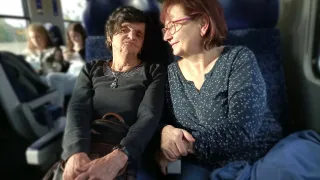 Zwei Frauen sitzen in einem Zug, eine mit einer Handtasche und die andere mit einer Brille. Sie scheinen beide in ein Gespräch vertieft zu sein.