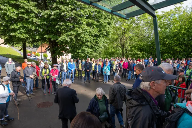 Eine große Gruppe von Menschen versammelt sich in einem Park. Einige tragen Hüte, Jacken und Brillen, während andere Stöcke tragen. Ein Mann in einem Anzug spricht zu der Menge, und Bäume umgeben das Gebiet.
