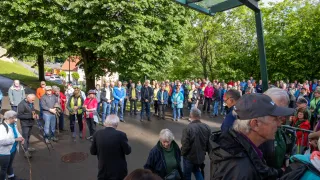 Eine große Gruppe von Menschen versammelt sich in einem Park. Einige tragen Hüte, Jacken und Brillen, während andere Stöcke tragen. Ein Mann in einem Anzug spricht zu der Menge, und Bäume umgeben das Gebiet.