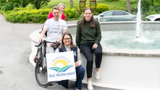 Drei Personen posieren neben einem Brunnen mit einem Schild für Bad Waltersdorf. Eine Frau hält ein Schild, ein Mann sitzt auf einem Fahrrad und eine weitere Frau sitzt hinter ihnen.