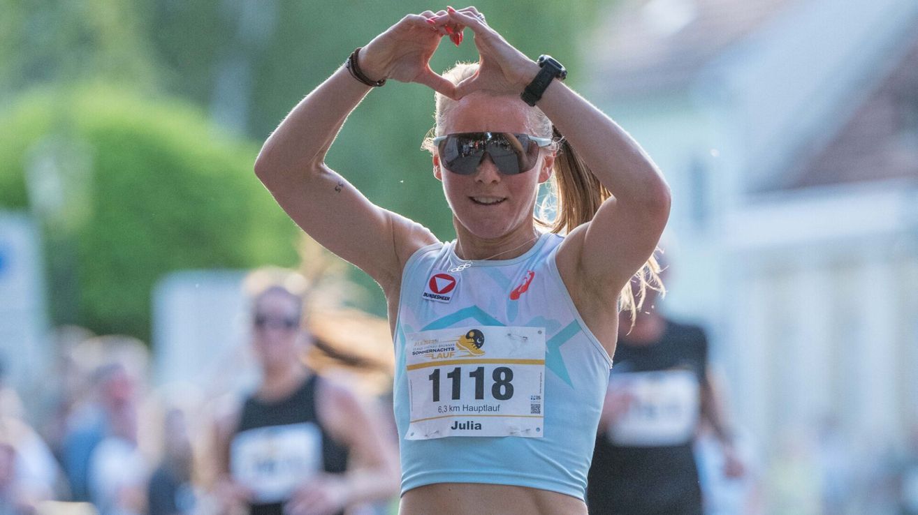 Eine Läuferin namens Julia, die eine Sonnenbrille und einen Nummernaufkleber 1118 trägt, bildet während eines Marathons mit ihren Händen ein Herz.