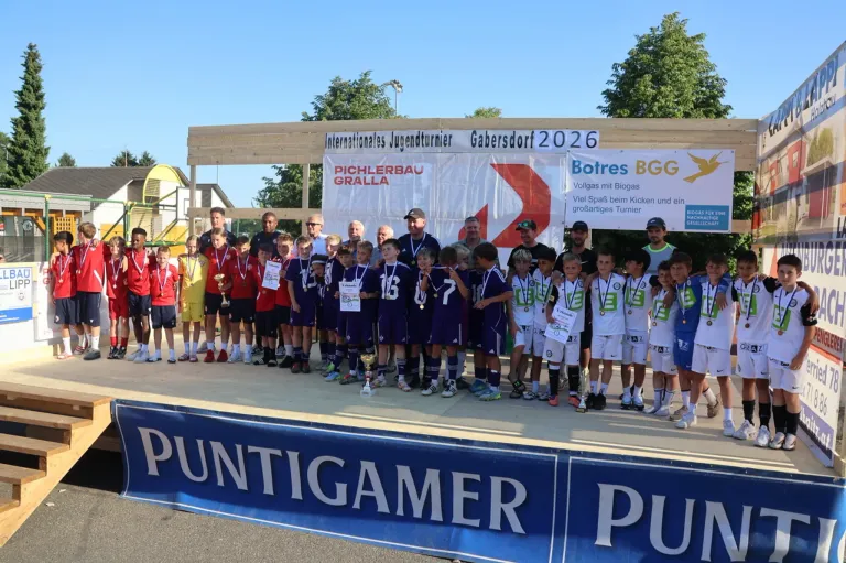 Eine Fußballmannschaft steht mit Medaillen, Trophäen und Erwachsenen auf der Bühne. Dahinter steht ein Banner mit der Aufschrift 'Internationales Jugendturnier Gabersdorf 2026'. Im Hintergrund sind Bäume und ein Gebäude zu sehen.