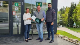 Drei Personen stehen vor einem Gebäude mit dem Schild 'Gemeindeamt Hollenthon'. Eine Frau in einer weißen Bluse und Jeans hält ein Zertifikat, während zwei Männer in grauen und blauen Jacken sie begleiten. Hinter ihnen sind Bäume und eine Bank zu sehen.