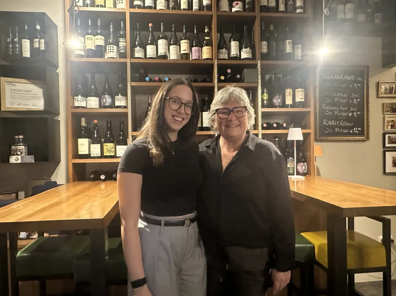 Zwei Frauen stehen in einem Weinlokal, eine mit Brille und Gürtel, die andere mit Halskette, mit einem Regal voller Flaschen hinter ihnen.