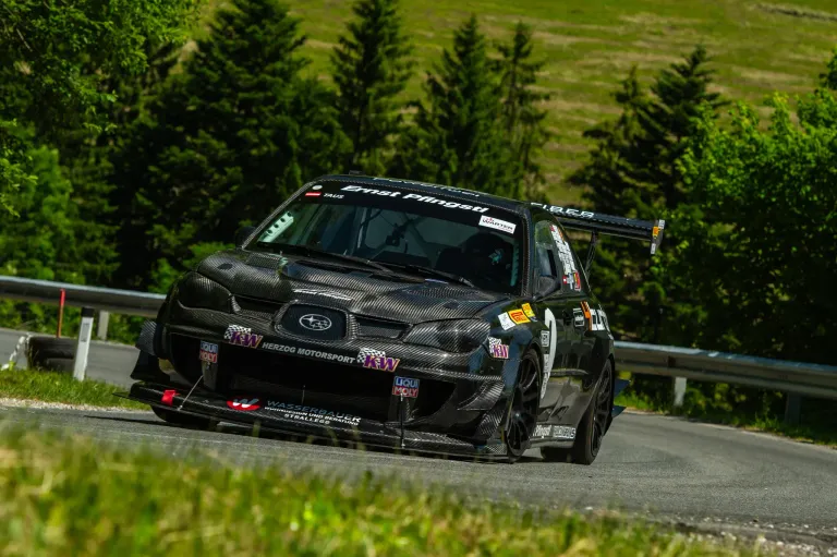 A black Subaru car with carbon fiber body panels is racing on a road with a grassy area. The car has the text Ernst Pingel on the hood.