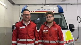 Zwei Männer in roten Rettungsdienstjacken posieren vor einem Krankenwagen. Einer trägt eine Brille und ein weisses Hemd, der andere hat einen Bart und eine rote Jacke mit 'Notfall-Sanitäter' darauf.
