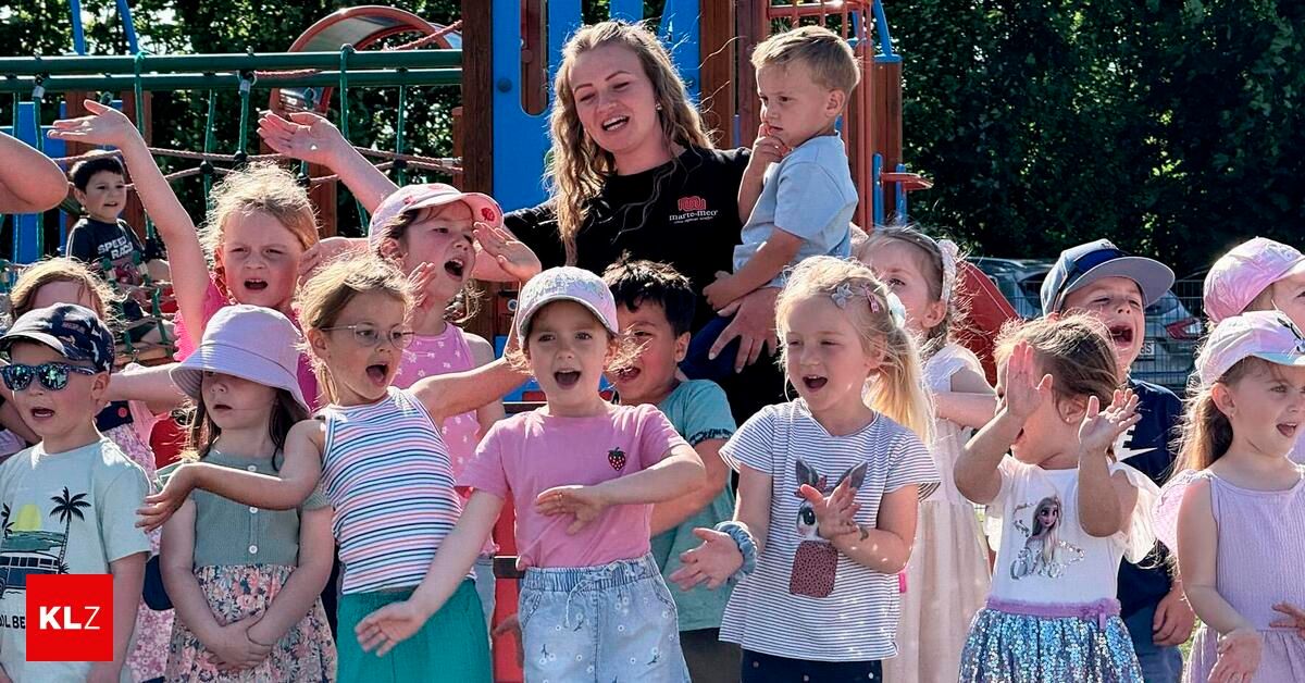Eine Gruppe von Kindern, einschließlich eines Babys, steht auf einem Spielplatz im Freien, wobei eine Frau ein Baby hält und eine andere Frau in der Front singt.