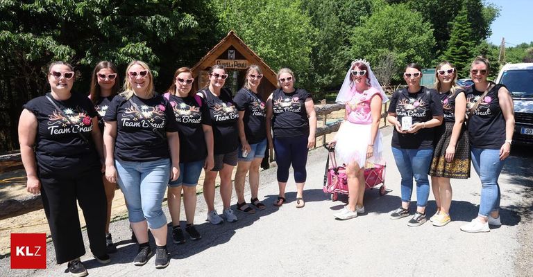 Eine Gruppe von Frauen in passenden T-Shirts posiert für ein Foto im Freien, eine in einem Hochzeitskleid, mit einer Holzhütte und Grün im Hintergrund.
