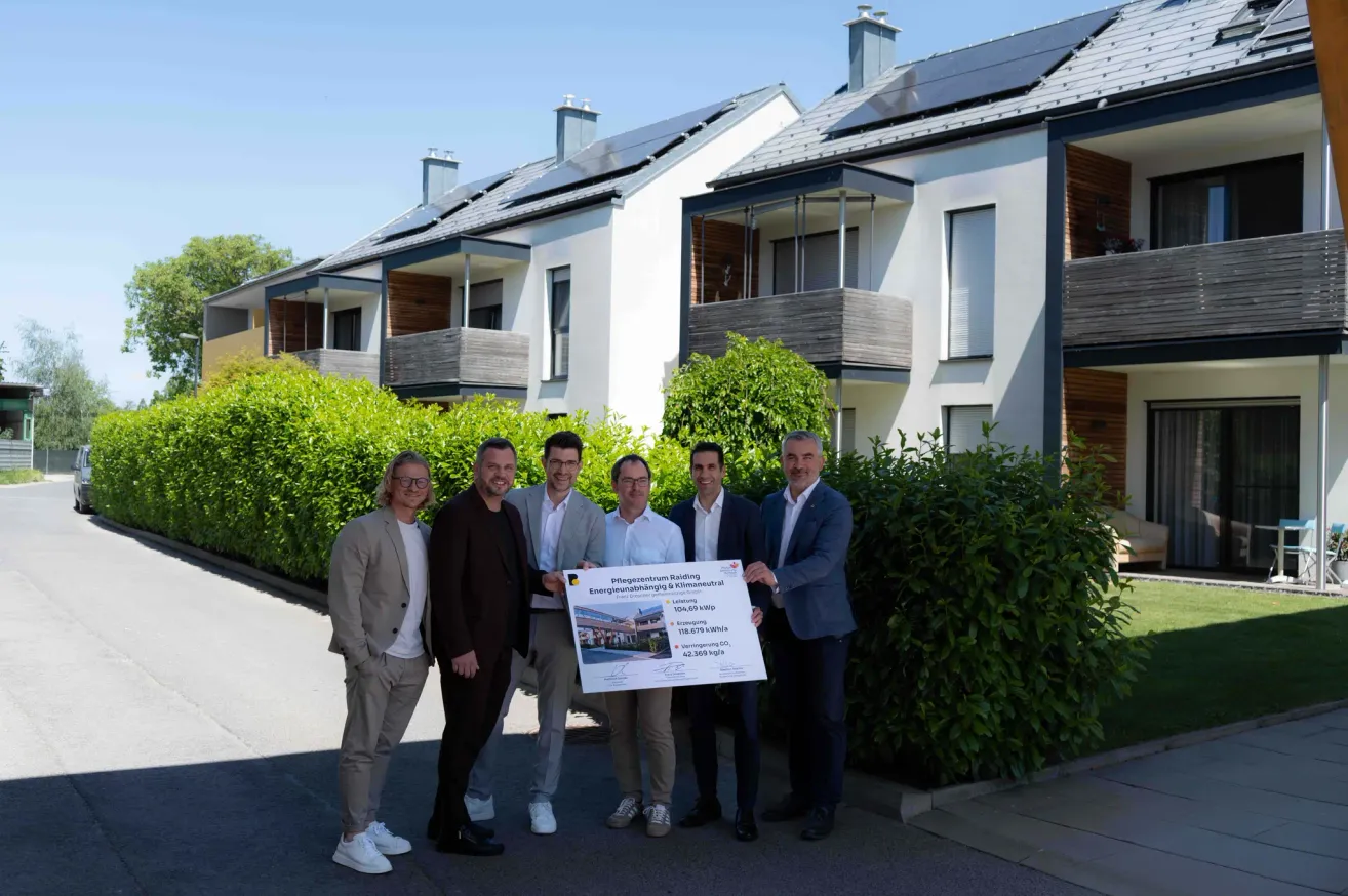 Fünf Männer stehen vor einem Haus mit Solarpaneelen und halten ein Zertifikat. Sie lächeln. Dahinter stehen Hecken und ein Haus.
