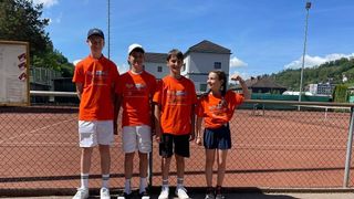 Fünf junge Personen in orangefarbenen T-Shirts posieren für ein Foto auf einem Tennisplatz. Sie stehen alle lächelnd vor einem Zaun. Dahinter befindet sich ein Haus mit Schornstein und Turm.