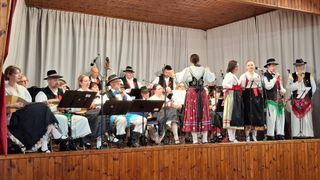 Eine Gruppe von Musikern in traditioneller Kleidung tritt auf der Bühne auf. Die Instrumente umfassen Gitarren, Cello und Geige. Das Publikum sitzt hinter ihnen auf Stühlen.