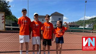 Vier Kinder in orangefarbenen Shirts stehen gemeinsam auf einem Tennisplatz und lächeln für ein Foto. Hinter ihnen steht ein Haus mit einem braunen Dach.