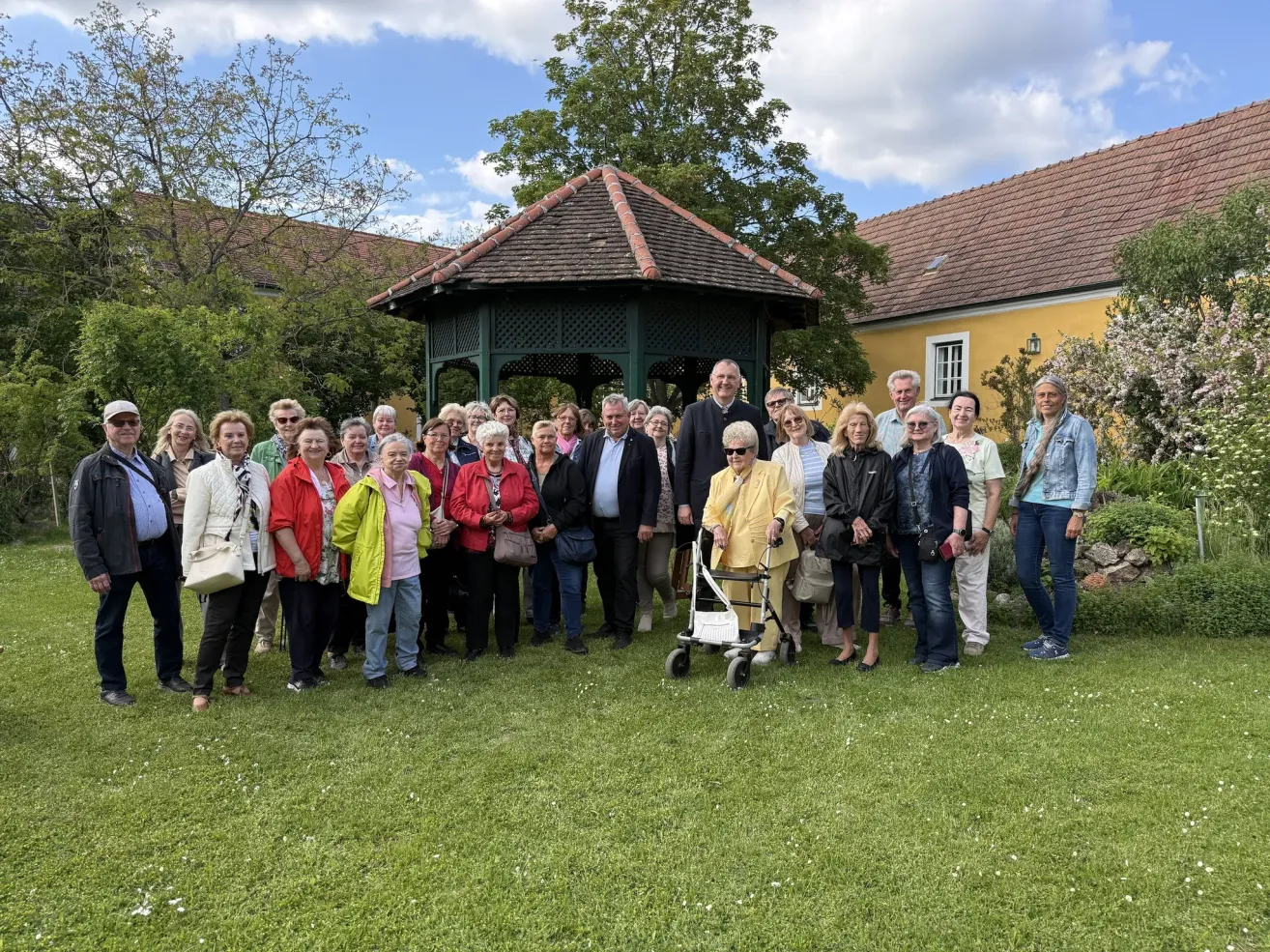 Eine große Gruppe von Menschen posiert für ein Foto in einem Garten mit einem Pavillon und einem gelben Haus im Hintergrund.
