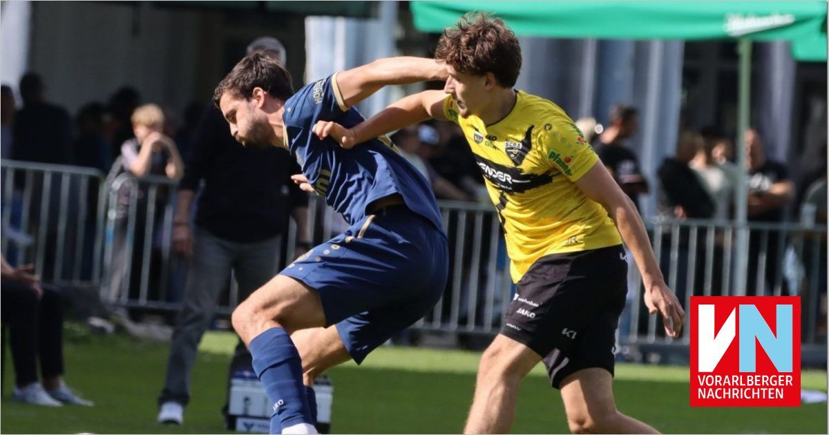 Zwei Fußballspieler, einer in Blau und einer in Gelb, sind in einem Duell auf dem Feld. Dahinter sind verschwommene Menschen und ein grünes Zelt.
