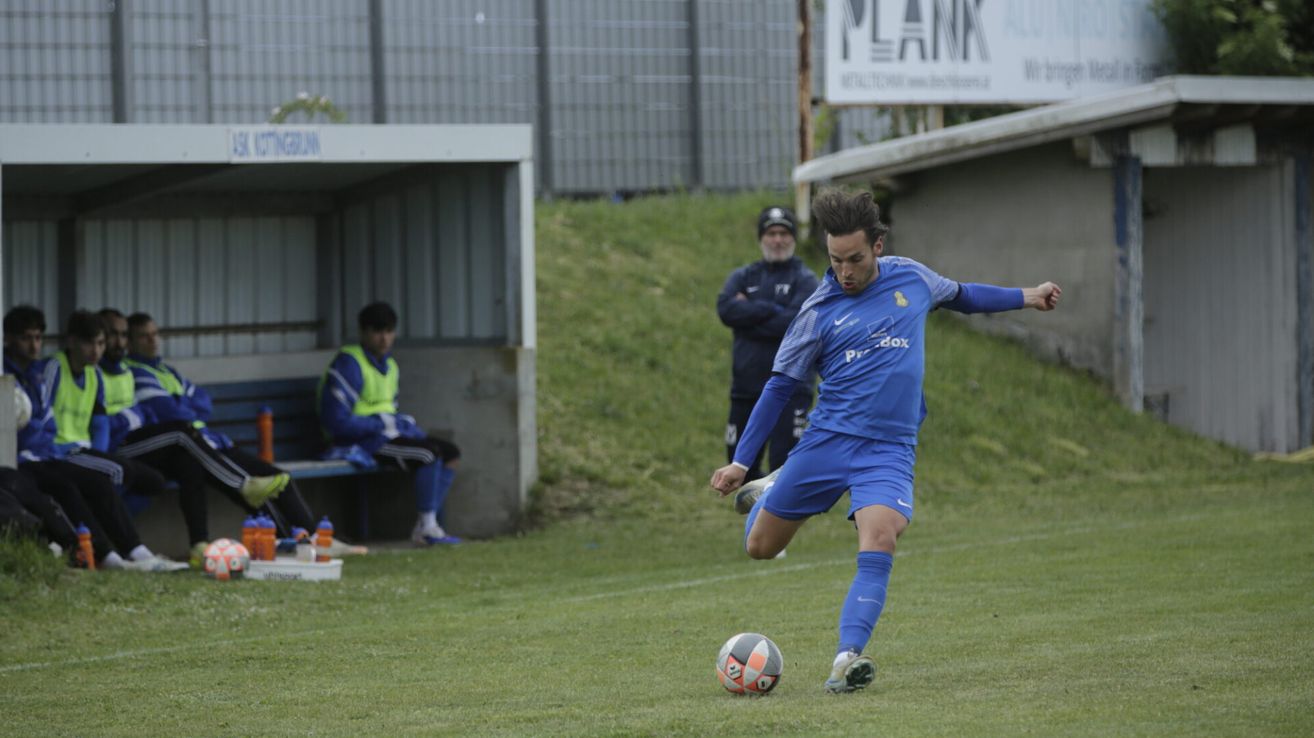 Ein Fußballer in Blau kickt einen Ball mit weißem und orangefarbenem Design auf dem Spielfeld. Dahinter beobachtet ein Mann und es gibt eine Bank mit einem sitzenden Mann. Im Hintergrund befindet sich ein Metallzaun.