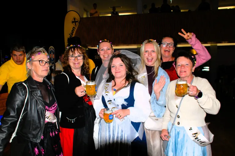 Group of women dressed in costumes holding mugs of beer, smiling and posing for a photo at a festival.