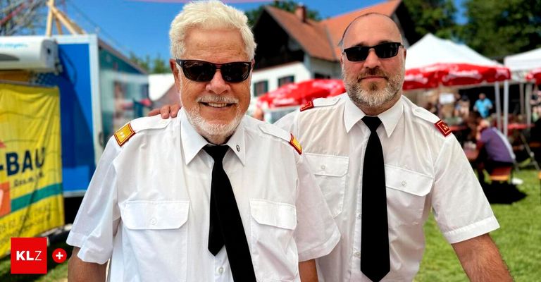 Zwei Männer in weißen Uniformen stehen draußen und lächeln. Einer hat einen weißen Bart, der andere eine Sonnenbrille. Sie stehen vor einem Gebäude mit roten Sonnenschirmen.