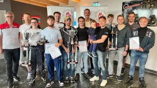Eine Gruppe von Männern und Frauen posiert mit Trophäen in einem Raum mit Holzbalken und einem gefliesten Boden.
