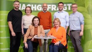 Fünf Personen stehen vor einem grünen Banner und lächeln. Zwei Frauen sitzen auf Stühlen, und ein Tisch mit Gläsern steht zwischen ihnen. Der Mann im orangefarbenen Pullover schaut direkt in die Kamera.