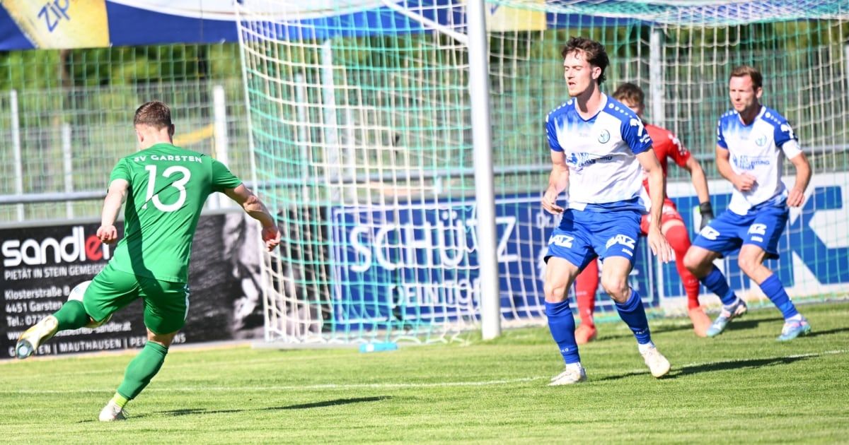 Fußballspieler rennen auf einem Feld; ein Spieler in einem blauen Trikot läuft auf das Tor zu. Ein anderer Spieler in einem grünen Trikot läuft auf ihn zu. Hinter ihnen steht ein Tor mit einem Netz.